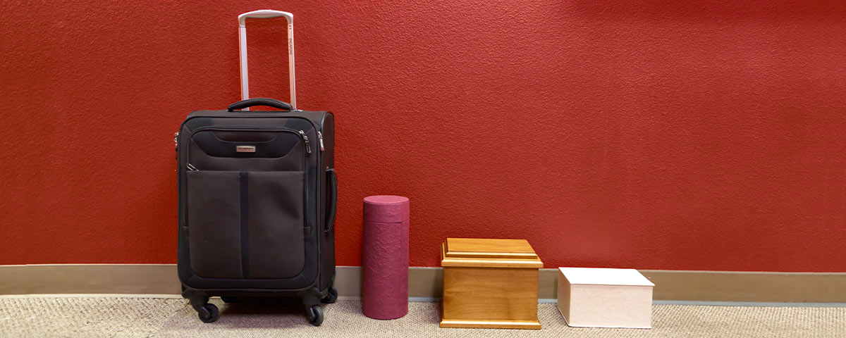 A black suitcase stands against the wall alongside a tube-shaped urn, a wooden urn, and a box-shaped urn.
