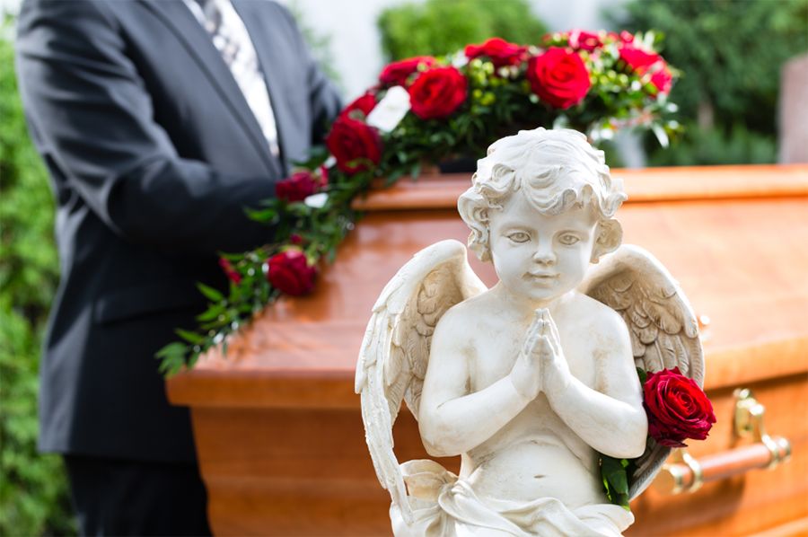 Marble statue of angel praying placed in front of brown casket and man in suit.