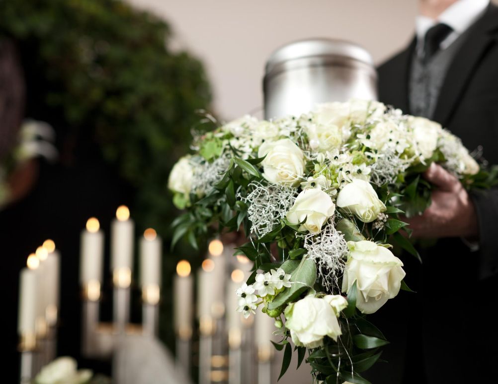 Man in funeral suit holding a silver cremation urn and a flower arrangement.