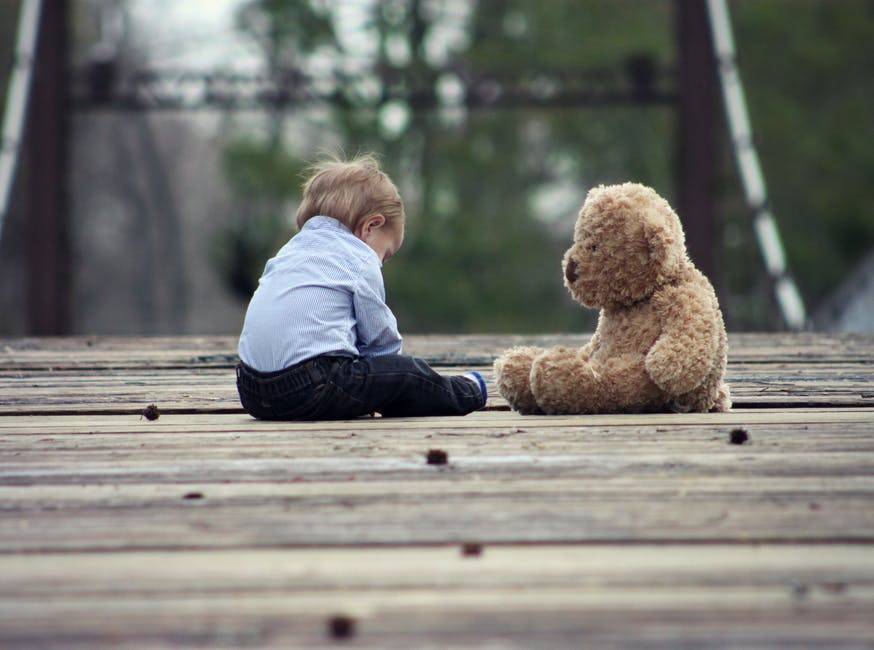 A toddler and a teddy bear sitting opposite each other on a bridge.