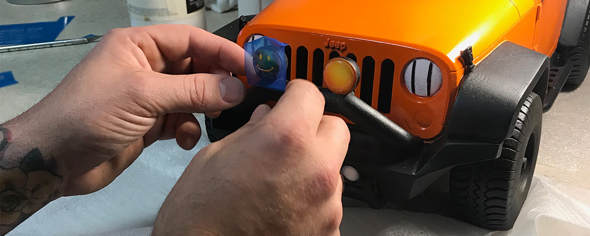 Hands apply a smiley face decal to the 3D-printed Jeep.