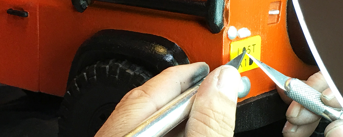 Hands hold two craft knives to the yellow L05T K1D license plate on the Land Rover urn.