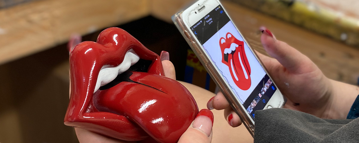 A freshly painted 3D-printed model of the tongue and lips design held in comparison to the original logo on a phone.