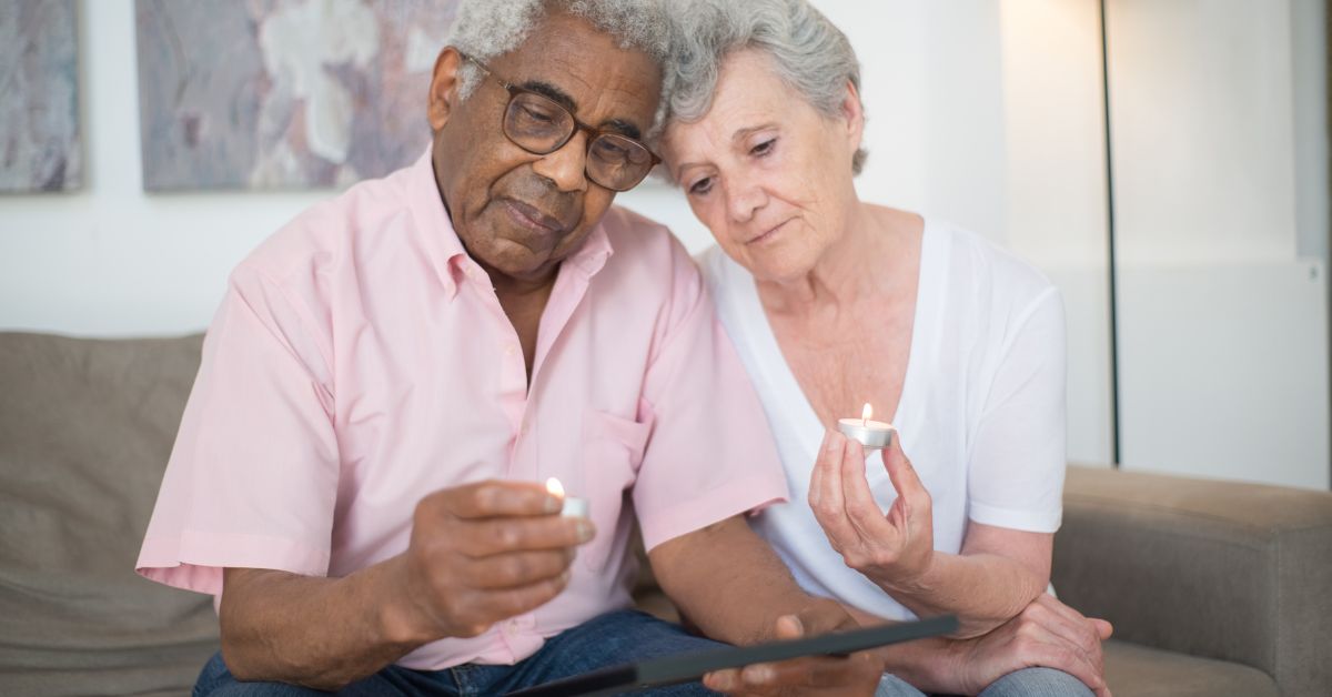 Elderly man and woman sitting together, each holding a burning candle in one hand while looking at a tablet.