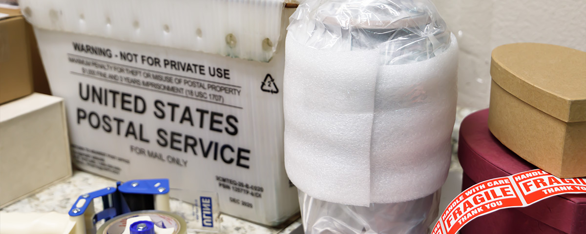 An urn is securely wrapped for shipping next to mailing supplies and a United States Postal Service bin.